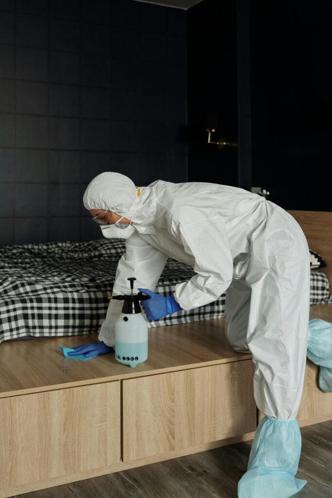 A worker in full protective gear disinfecting a bedroom with a spray bottle and cloth.